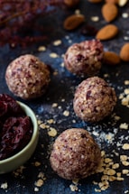 Round energy balls with bits of oats and dried fruit are displayed on a dark surface. A small bowl filled with dried cranberries is nearby, surrounded by scattered oats and almonds.