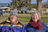 An older couple is sitting on a bench outdoors, smiling at the camera. The man is wearing glasses, a gray beanie with a light on it, and a blue and yellow jacket. The woman has a fluffy scarf and a blue fleece jacket. In the background, there are trees, parked cars, including an RV, and buildings.