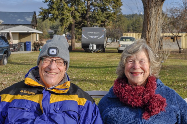 An older couple is sitting on a bench outdoors, smiling at the camera. The man is wearing glasses, a gray beanie with a light on it, and a blue and yellow jacket. The woman has a fluffy scarf and a blue fleece jacket. In the background, there are trees, parked cars, including an RV, and buildings.