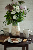 A rustic wooden table set for breakfast with colorful ceramic mugs, plates, and a vase of fresh flowers.