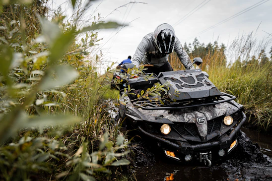 A person wearing a helmet navigates an all-terrain vehicle (ATV) through a muddy, off-road trail surrounded by tall grass and foliage. The ATV's front wheels are partially submerged, and water can be seen splashing around. The background is filled with more vegetation and hints of a forest.