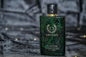 A green rectangular perfume bottle with a golden cap, featuring droplets of water on its surface, placed against a dark, possibly velvet-like background. The bottle has a sophisticated design with white text and a laurel wreath emblem.