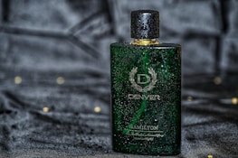 A green rectangular perfume bottle with a golden cap, featuring droplets of water on its surface, placed against a dark, possibly velvet-like background. The bottle has a sophisticated design with white text and a laurel wreath emblem.