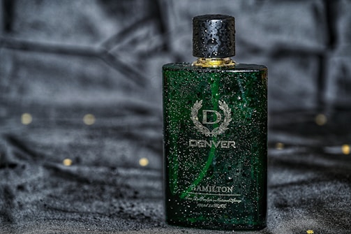 A green rectangular perfume bottle with a golden cap, featuring droplets of water on its surface, placed against a dark, possibly velvet-like background. The bottle has a sophisticated design with white text and a laurel wreath emblem.