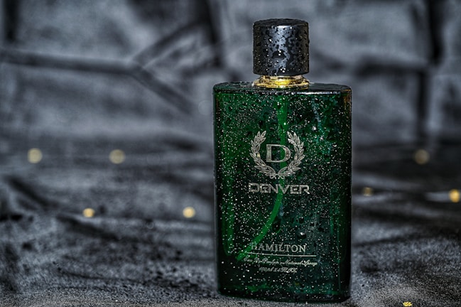 A green rectangular perfume bottle with a golden cap, featuring droplets of water on its surface, placed against a dark, possibly velvet-like background. The bottle has a sophisticated design with white text and a laurel wreath emblem.