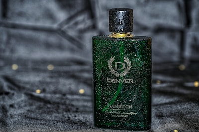 A green rectangular perfume bottle with a golden cap, featuring droplets of water on its surface, placed against a dark, possibly velvet-like background. The bottle has a sophisticated design with white text and a laurel wreath emblem.