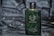 A green rectangular perfume bottle with a golden cap, featuring droplets of water on its surface, placed against a dark, possibly velvet-like background. The bottle has a sophisticated design with white text and a laurel wreath emblem.