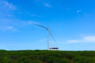 A large wind turbine towers over a green landscape with a white van parked nearby. The sky is clear with a bright blue color, contributing to the serene atmosphere.