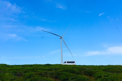 A large wind turbine towers over a green landscape with a white van parked nearby. The sky is clear with a bright blue color, contributing to the serene atmosphere.