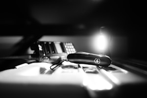 A monochrome image featuring a Swiss Army knife prominently placed in the foreground on a reflective surface. In the background, to the left, there's a portable radio with an antenna and various buttons. The lighting is dramatic, with a bright light source creating high contrast and deep shadows.