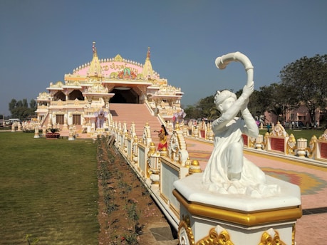 A detailed temple facade with intricate designs highlighted by golden accents. In the foreground, a white statue of a person holding a curved instrument draws attention. The temple is surrounded by a neatly kept green lawn and pathways with decorative elements. The sky is clear and blue, adding to the peaceful ambiance.