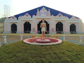 An intricately designed tent structure with colorful decorations and ornate patterns is visible. A statue of a historically styled figure is positioned prominently on a circular platform in front of the building. The structure is adorned with arched entrances and decorative motifs. Chairs are arranged inside the tent, suggesting a venue prepared for a gathering or event. The ground is grassy, and some decorative pillars flank the platform.