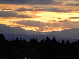 The image captures a dramatic sunset over a mountainous landscape. The sky is filled with clouds, creating a mix of dark and light areas, with a warm, fiery glow from the setting sun. The silhouette of trees and rooftops can be seen in the foreground, adding depth to the scene.