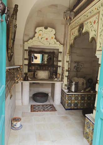 A traditional room with ornate Islamic architectural elements, featuring intricately patterned tiles on the lower walls and ceiling. The space includes an old stone basin on a table with decorative woodwork and mirrors. There are also traditional metal utensils and decorative pottery placed around. The floor is tiled, and the overall decor suggests a historical or cultural setting.