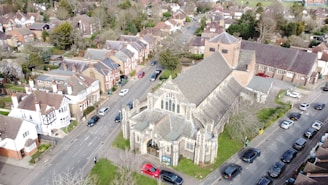 An aerial view of a residential area featuring a large stone church with a pitched roof and stained glass windows. Surrounding the church are rows of houses with a mixture of traditional and modern architectural styles. Several cars are parked along the street, and there are patches of greenery and trees throughout the neighborhood. The scene has a suburban atmosphere with neatly arranged streets and houses.