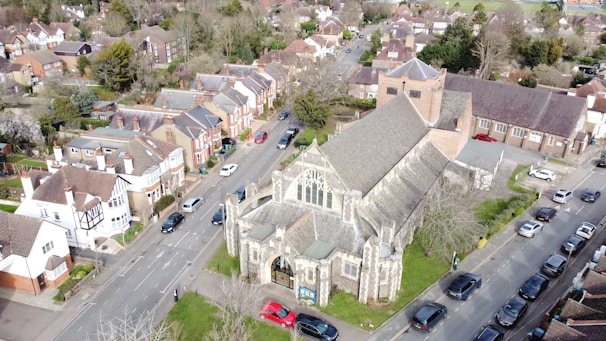 An aerial view of a residential area featuring a large stone church with a pitched roof and stained glass windows. Surrounding the church are rows of houses with a mixture of traditional and modern architectural styles. Several cars are parked along the street, and there are patches of greenery and trees throughout the neighborhood. The scene has a suburban atmosphere with neatly arranged streets and houses.