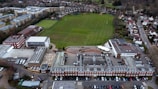 An aerial view of a large complex featuring a traditional red brick building with a gray roof, adjacent to a spacious green field marked for sports. Surrounding the central structure are several modern buildings with varied architectural styles, including flat roofs and a red and white design. Nearby, residential housing and other urban infrastructure are visible, providing a sense of a well-organized suburban environment.