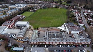 An aerial view of a large complex featuring a traditional red brick building with a gray roof, adjacent to a spacious green field marked for sports. Surrounding the central structure are several modern buildings with varied architectural styles, including flat roofs and a red and white design. Nearby, residential housing and other urban infrastructure are visible, providing a sense of a well-organized suburban environment.