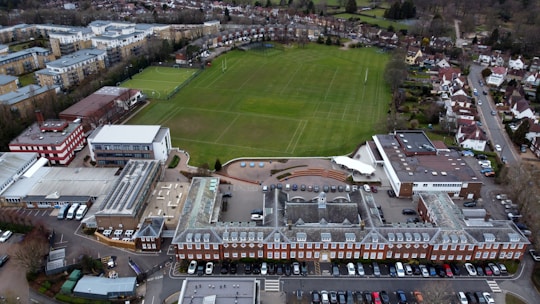 An aerial view of a large complex featuring a traditional red brick building with a gray roof, adjacent to a spacious green field marked for sports. Surrounding the central structure are several modern buildings with varied architectural styles, including flat roofs and a red and white design. Nearby, residential housing and other urban infrastructure are visible, providing a sense of a well-organized suburban environment.