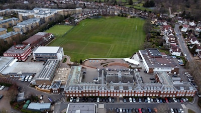 An aerial view of a large complex featuring a traditional red brick building with a gray roof, adjacent to a spacious green field marked for sports. Surrounding the central structure are several modern buildings with varied architectural styles, including flat roofs and a red and white design. Nearby, residential housing and other urban infrastructure are visible, providing a sense of a well-organized suburban environment.