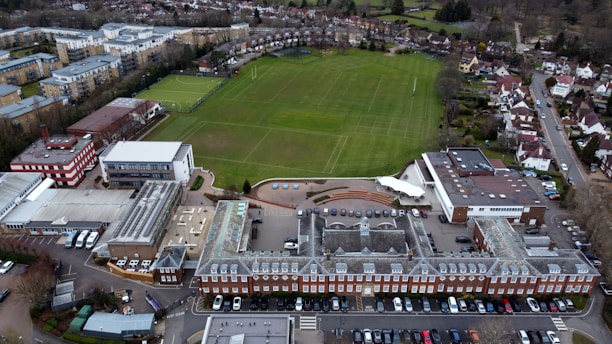 An aerial view of a large complex featuring a traditional red brick building with a gray roof, adjacent to a spacious green field marked for sports. Surrounding the central structure are several modern buildings with varied architectural styles, including flat roofs and a red and white design. Nearby, residential housing and other urban infrastructure are visible, providing a sense of a well-organized suburban environment.