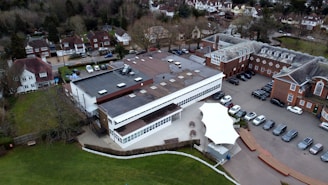 An aerial view of a large building complex surrounded by a mix of residential homes and a parking area. The central structure appears modern with a flat roof and numerous skylights, next to older, more traditional red brick buildings. Cars are parked in rows along a paved area, and there is a green lawn on the left side. Trees and residential houses are visible in the background.