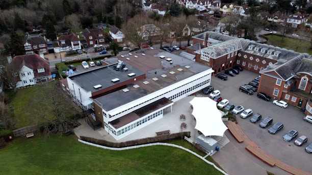 An aerial view of a large building complex surrounded by a mix of residential homes and a parking area. The central structure appears modern with a flat roof and numerous skylights, next to older, more traditional red brick buildings. Cars are parked in rows along a paved area, and there is a green lawn on the left side. Trees and residential houses are visible in the background.