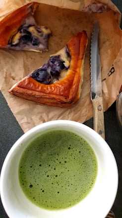 A cup of frothy green matcha tea is placed next to two slices of blueberry-topped pastry on a piece of brown parchment paper. A serrated knife with a wooden handle rests near the pastry.