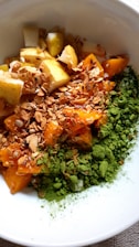 A bowl filled with chopped pears, bright orange apricots, granola, and a generous portion of green matcha powder, all arranged artistically.