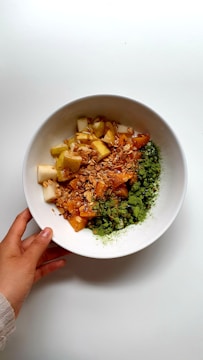 A white bowl filled with a mixture of chopped fruits, granola, and a green powder is held by a hand over a white background. The fruits appear to be diced with an assortment of textures and colors, including yellow and orange shades. The granola adds a crunchy texture, while the green powder, possibly matcha or crushed nuts, covers part of the ingredients.