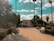 A pathway surrounded by trees and benches leads towards a gate. Palm trees and shrubs line the path, and there is a building visible to the right. The sky is partly cloudy with patches of blue.