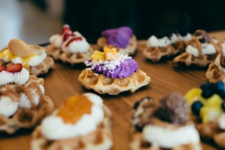 A selection of intricately decorated waffles is arranged on a wooden table. Each waffle is uniquely topped with colorful and varied toppings, such as purple icing, whipped cream, fruits like blueberries and strawberries, and other sweet garnishes.
