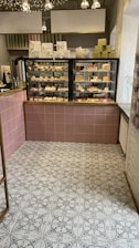 A cozy café interior featuring a display case filled with various pastries and desserts. Above the case, there are signs in a foreign language detailing menu options. The floor has a patterned tile design, and a selection of gift boxes is arranged on top of the counter.