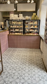 A cozy café interior featuring a display case filled with various pastries and desserts. Above the case, there are signs in a foreign language detailing menu options. The floor has a patterned tile design, and a selection of gift boxes is arranged on top of the counter.