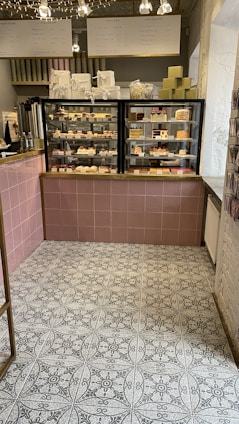 A cozy café interior featuring a display case filled with various pastries and desserts. Above the case, there are signs in a foreign language detailing menu options. The floor has a patterned tile design, and a selection of gift boxes is arranged on top of the counter.