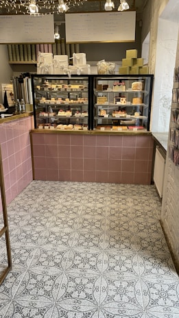 A cozy café interior featuring a display case filled with various pastries and desserts. Above the case, there are signs in a foreign language detailing menu options. The floor has a patterned tile design, and a selection of gift boxes is arranged on top of the counter.