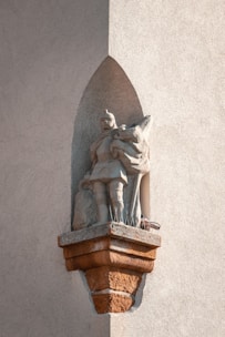 A stone statue of a medieval knight placed in an alcove on a rough-textured wall. The knight is wearing armor and holds a shield, with a distinct shadow cast by the sunlight. The base of the statue has anti-bird spikes.