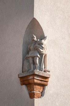 A stone statue of a medieval knight placed in an alcove on a rough-textured wall. The knight is wearing armor and holds a shield, with a distinct shadow cast by the sunlight. The base of the statue has anti-bird spikes.