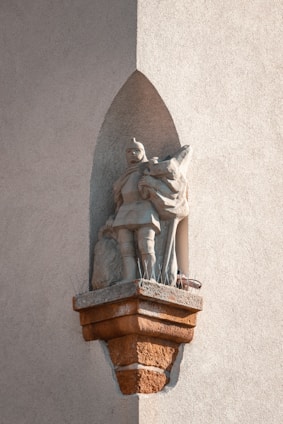 A stone statue of a medieval knight placed in an alcove on a rough-textured wall. The knight is wearing armor and holds a shield, with a distinct shadow cast by the sunlight. The base of the statue has anti-bird spikes.