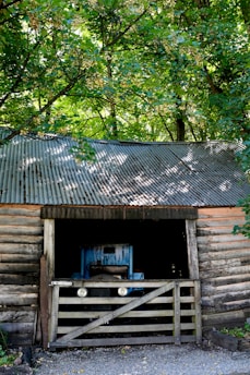 A rustic wooden shed surrounded by lush green trees, with a corrugated metal roof. Inside the barn, a blue tractor is partially visible, shielded by a wooden gate.