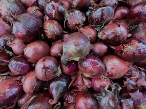 A close-up view of a large pile of red onions, displaying various sizes and shapes. The onions have a rich, deep purple outer layer with visible dry, papery skin peelings. Some have small roots still attached, adding to the natural texture.