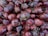 A close-up view of a large pile of red onions, displaying various sizes and shapes. The onions have a rich, deep purple outer layer with visible dry, papery skin peelings. Some have small roots still attached, adding to the natural texture.