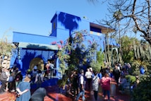 A vibrant blue building with intricate lattice work and floral decorations is surrounded by lush greenery and cactus plants. A group of people are gathered outside, some standing in line, while others explore the garden area. The scene is set under a clear blue sky, providing a bright and lively atmosphere.