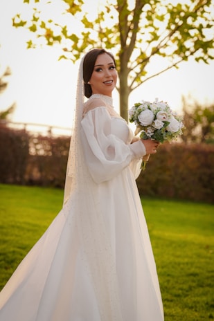 Bride in an elegant wedding dress smiling while holding a bouquet at a sunny outdoor venue.
