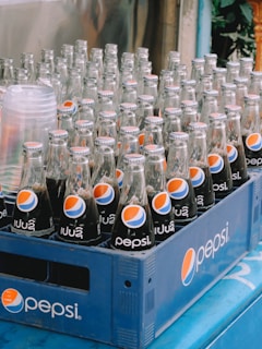 Several glass bottles of Pepsi are neatly arranged in a blue plastic crate. The bottles have a classic design with the Pepsi logo prominently displayed. The crate is placed on a blue surface, and next to it are some clear plastic cups.