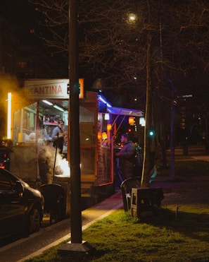 A street food vendor operates a brightly lit stand at night. The vendor is serving customers under neon lights that create a warm and inviting atmosphere. Smoke rises from the cooking grill, adding a dynamic element to the scene. Nearby, a person waits by a parked car as another customer is attended to, all set against the backdrop of a city street.