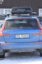 A blue Volvo XC60 T8 with a Norwegian license plate is parked outdoors. The vehicle has a roof box labeled 'Packline' mounted on top. The car and surrounding ground show traces of snow, suggesting a cold or wintery environment. The building in the background has a wooden façade with visible windows.