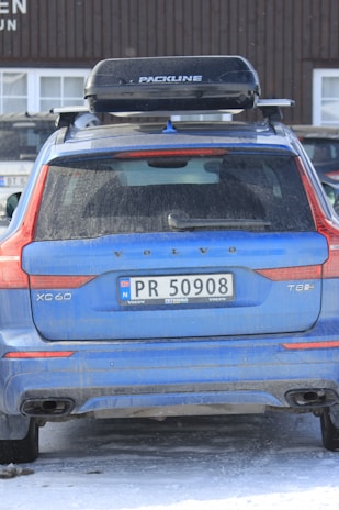 A blue Volvo XC60 T8 with a Norwegian license plate is parked outdoors. The vehicle has a roof box labeled 'Packline' mounted on top. The car and surrounding ground show traces of snow, suggesting a cold or wintery environment. The building in the background has a wooden façade with visible windows.