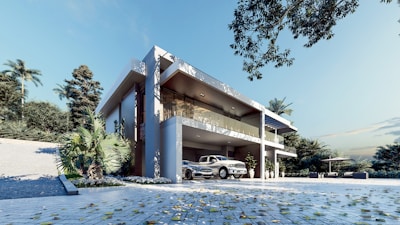 A modern two-story house with clean, angular architecture surrounded by lush greenery. The building features large glass windows and a spacious driveway with a vintage car parked in front. The setting includes a clear blue sky and scattered leaves on the ground, suggesting a breezy and serene environment.