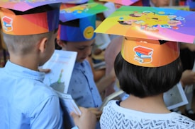 Children wearing colorful paper hats with educational-themed designs are viewing illustrated books. The hats are decorated with images of books and cartoon faces, suggesting a celebration or educational event.